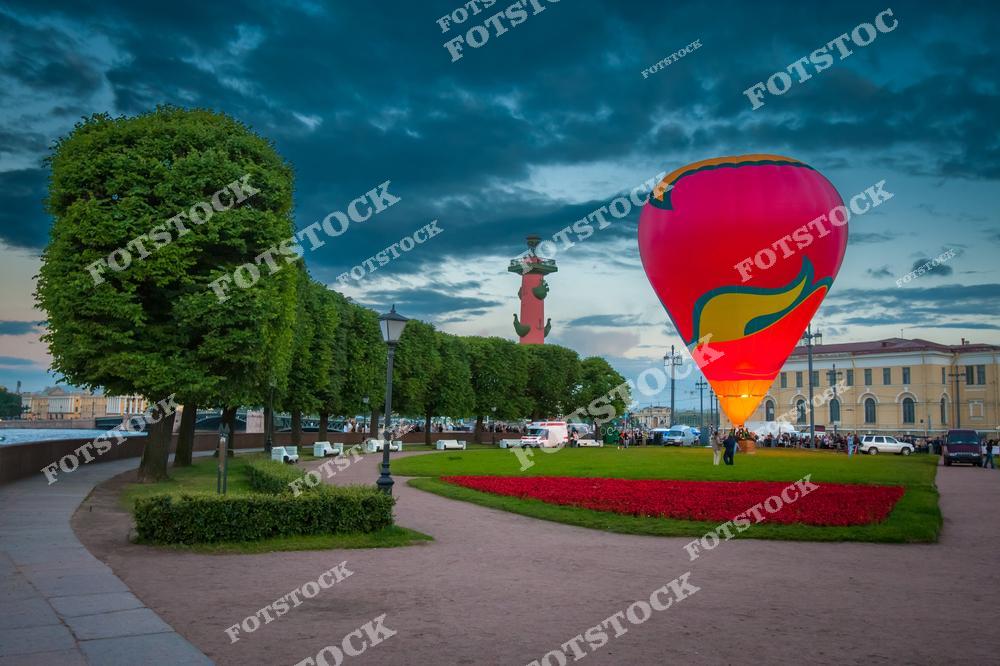 На фотографии изображен воздушный шар, поднимающийся в вечернее небо рядом с набережной в Санкт-Петербурге. Вдали видны Ростральные колонны на Стрелке Васильевского острова, одно из самых известных мест города. Зеленая аллея и цветочные клумбы создают живописный фон для этого яркого зрелища. Вечерняя атмосфера и спокойное освещение набережной делают картину особенно живописной.